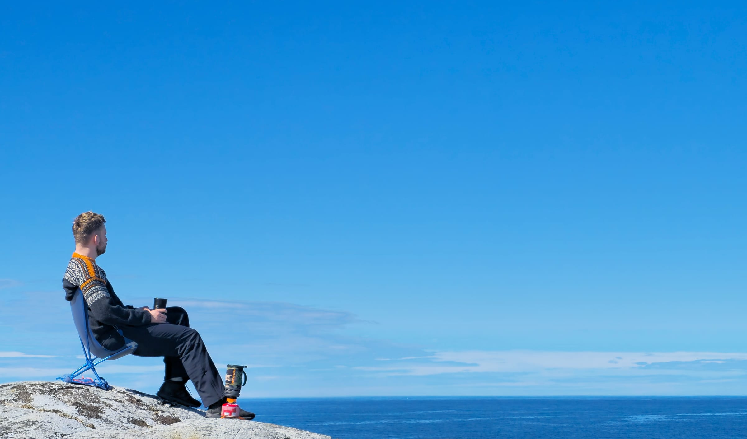 Hiker in a Norwegian Marius sweater sitting in a camp chair on coastal rocks with a coffee cup and camp stove
